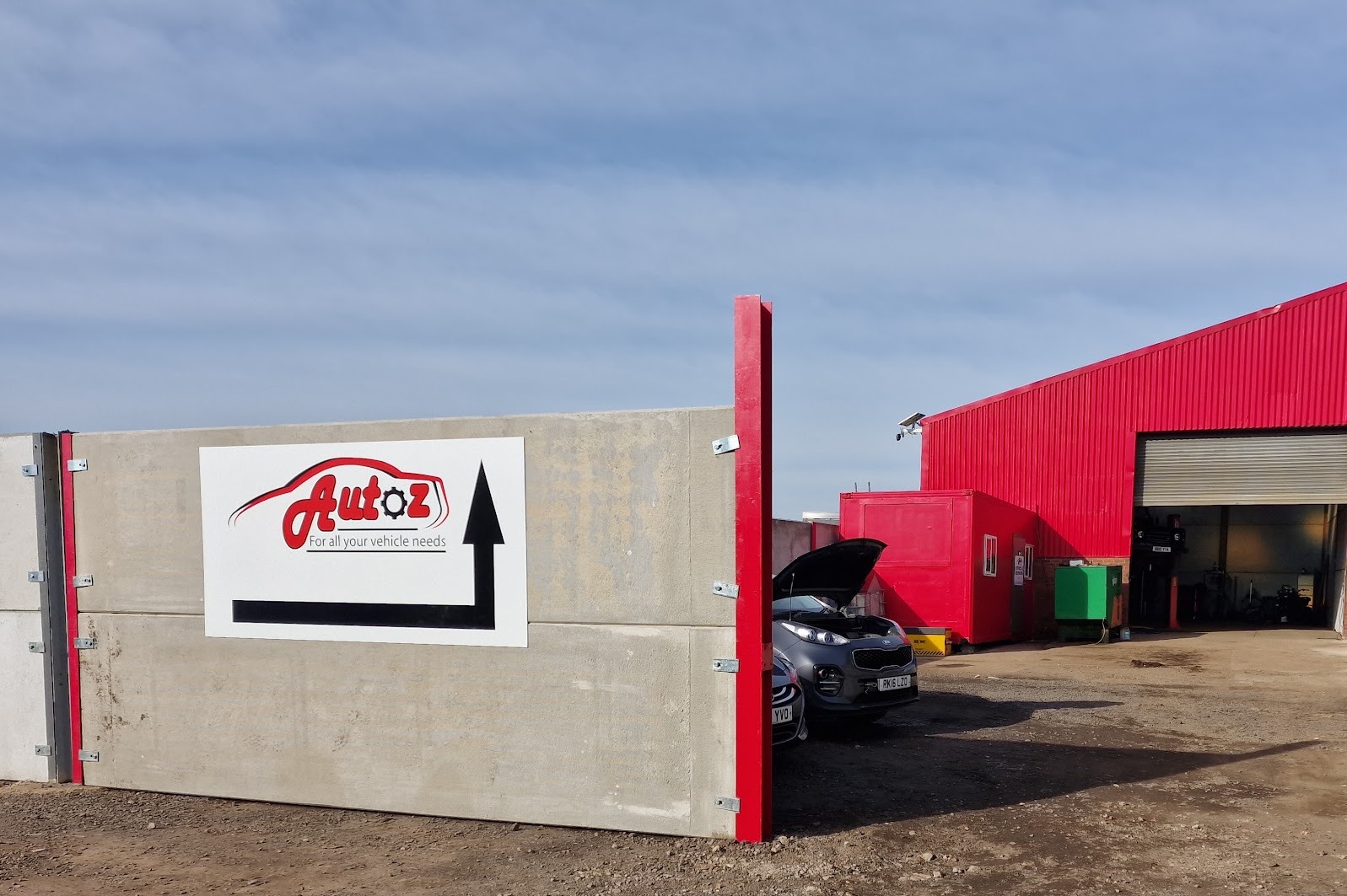 The entrance to Autoz: a concrete wall with the white Autoz sign and an arrow pointing into the yard, and the red workshop beyond