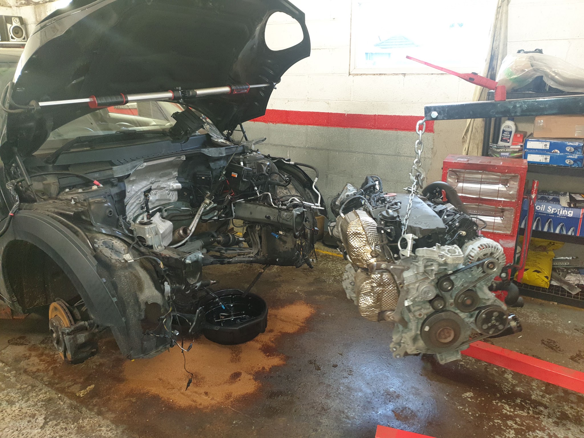A car on the workshop floor with its engine out and the engine hanging from a hoist beside it